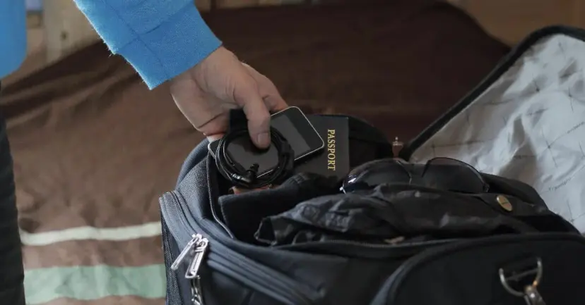 A hand packs a passport and a phone into a black travel bag.
