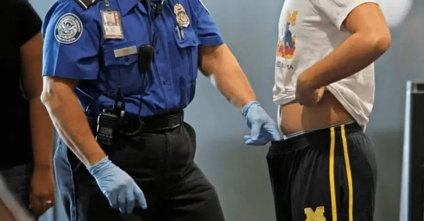 Airport security officer in blue gloves patting down a man who is lifting his shirt at a checkpoint.