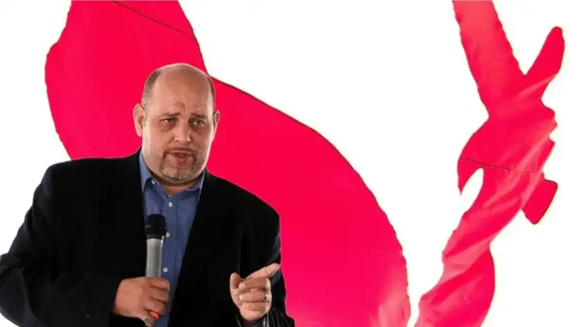 Bald man in a dark jacket speaking into a handheld microphone in front of a red and white backdrop.