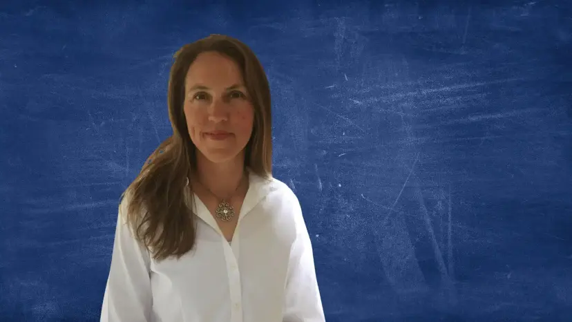 Angela Wetuski, a woman with long brown hair in a white blouse, in front of a blue chalkboard background.