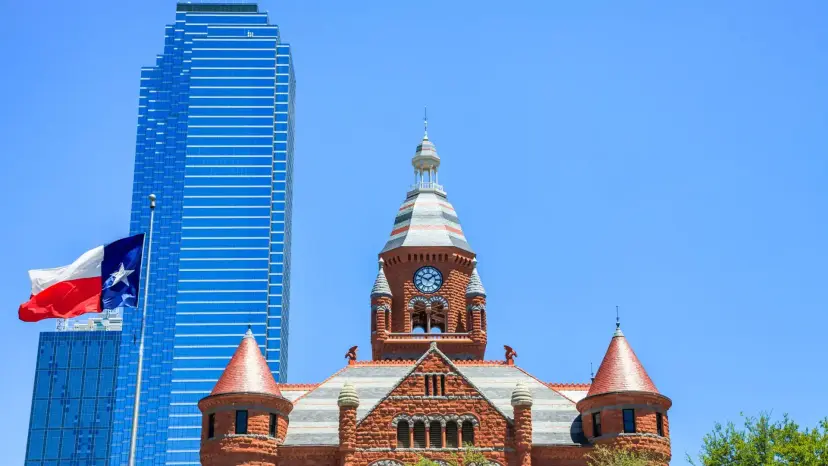 A Texas flag beside a blue glass skyscraper and the red stone Old Red Courthouse in downtown Dallas.