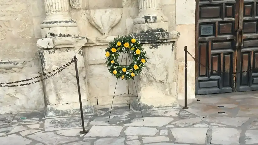 A wreath of yellow roses on a stand in front of the Alamo's carved stone facade and wooden doors.