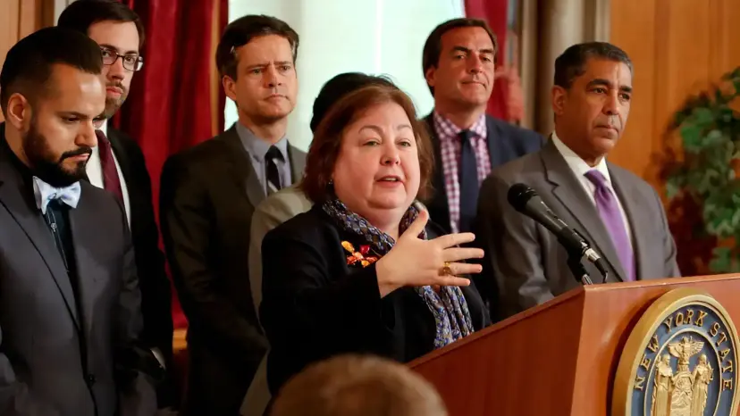 New York State Senator Liz Krueger speaks at a New York State podium, surrounded by men in suits.
