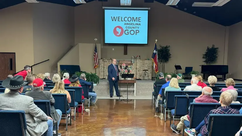 Man speaks at a lectern to a seated audience under a screen reading 'Welcome Angelina County GOP.'