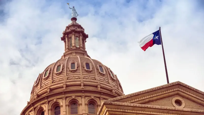 The Texas Capitol dome in Austin with a Texas flag flying against a cloudy sky.
