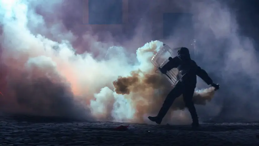 A person in dark gear carrying a riot shield walks through thick clouds of tear gas at night.