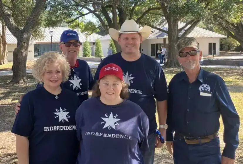 Five TNM members in 'Texas Independence' shirts and caps pose together under oak trees on a sunny day.