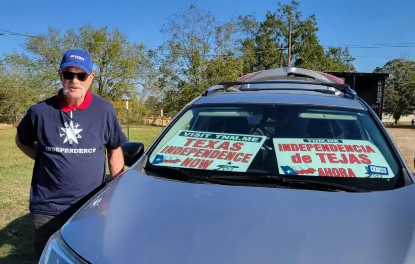 A man in a TEXIT cap stands by a car with windshield signs reading 'Texas Independence Now' and Spanish text.