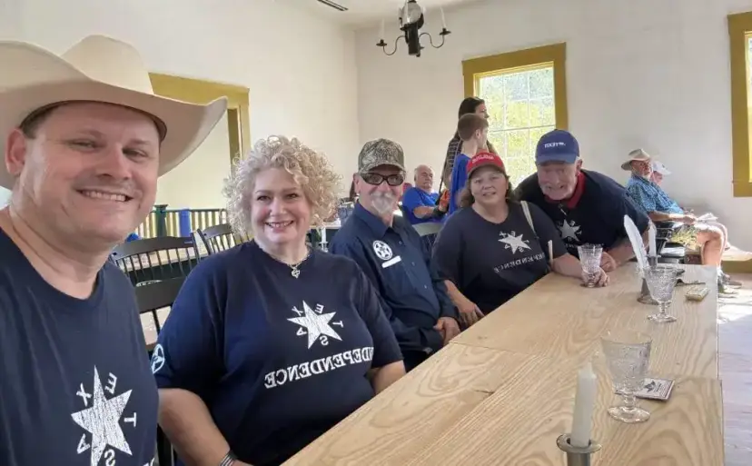 Smiling TNM members in 'Texas Independence' shirts sit at a long wooden table inside a historic building.