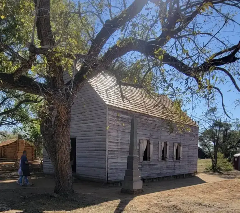 Independence Hall, a weathered wooden building with a stone monument in front, under a large tree at Washington-on-the-Brazos.