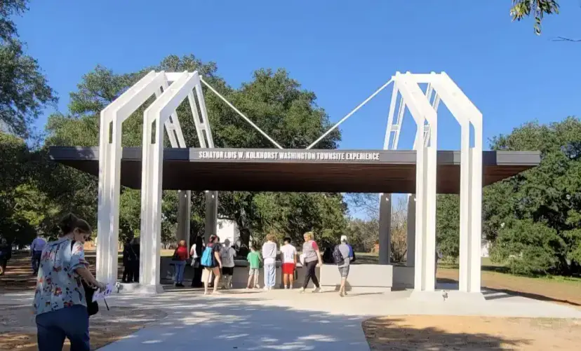 Visitors walk under a white-framed pavilion signed 'Senator Lois W. Kolkhorst Washington Townsite Experience.'