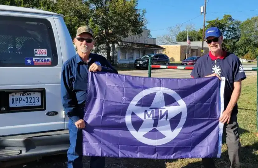 Two men hold a purple TNM flag beside a white van with Texas plates and a 'TEXIT Now' sticker.