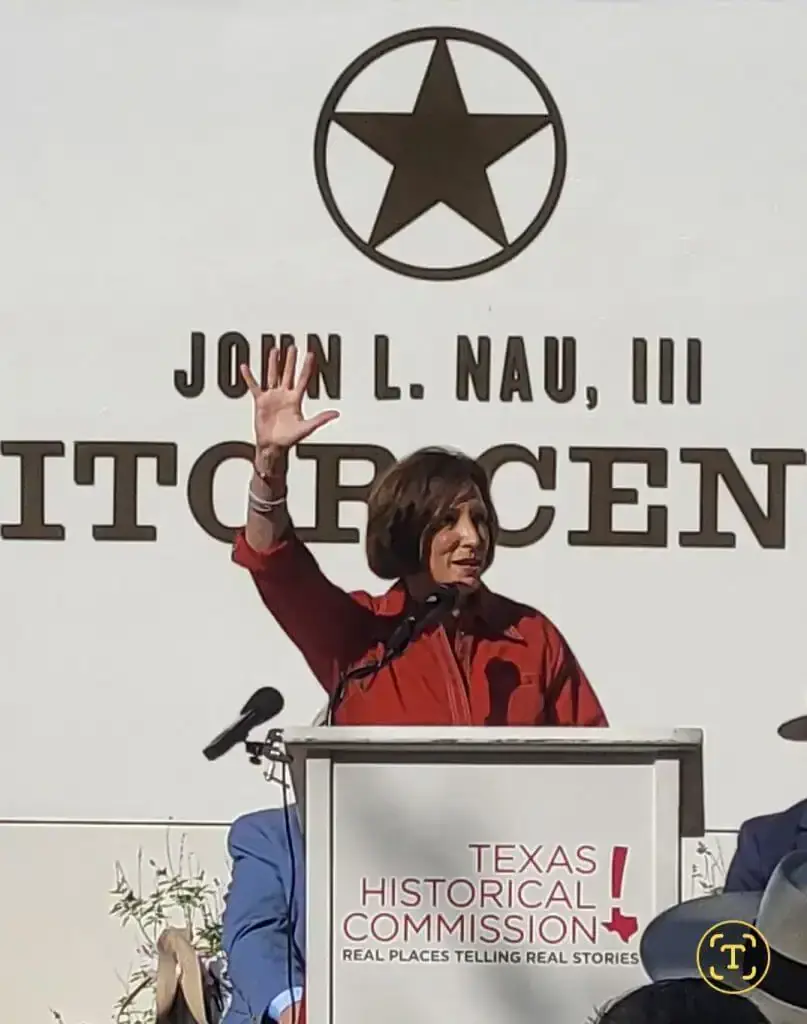 A woman in red waves from a Texas Historical Commission podium below the John L. Nau III visitor center sign.