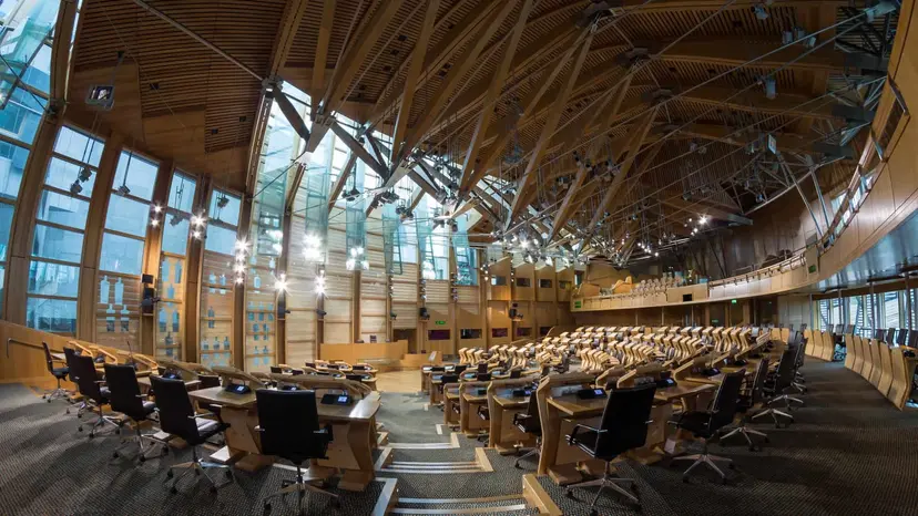 A debating chamber with curved benches and desks, empty, lit from a high glass ceiling.