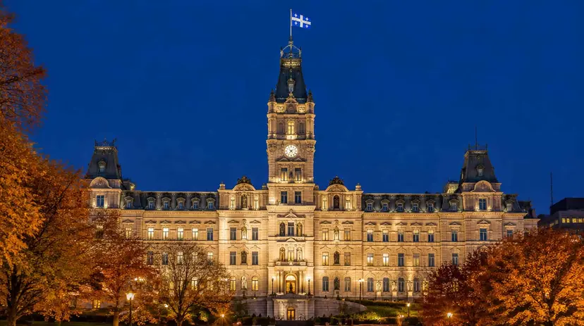 Quebec's Parliament Building in Quebec City lit at night, the Fleurdelisé flying from its clock tower, framed by autumn trees.
