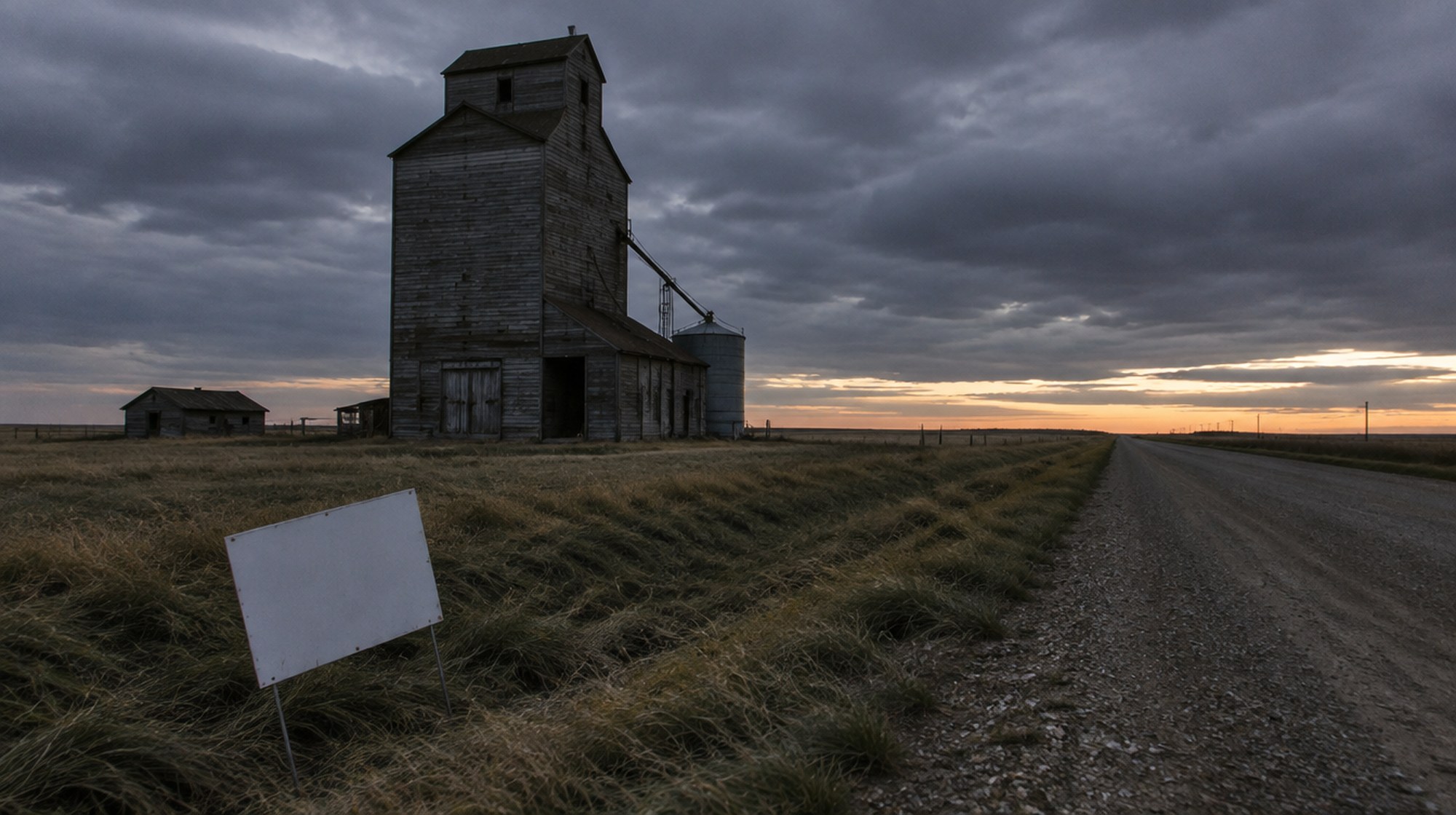 A weathered grain elevator beside a gravel road on the Alberta prairie at dusk, with a blank lawn sign in the ditch.