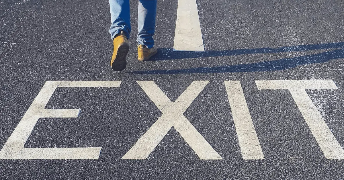 A person in work boots walks over the word 'Exit' painted on asphalt.