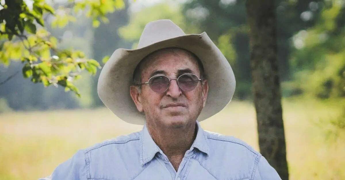 An older man in a cowboy hat, tinted glasses, and a denim shirt stands in a field.