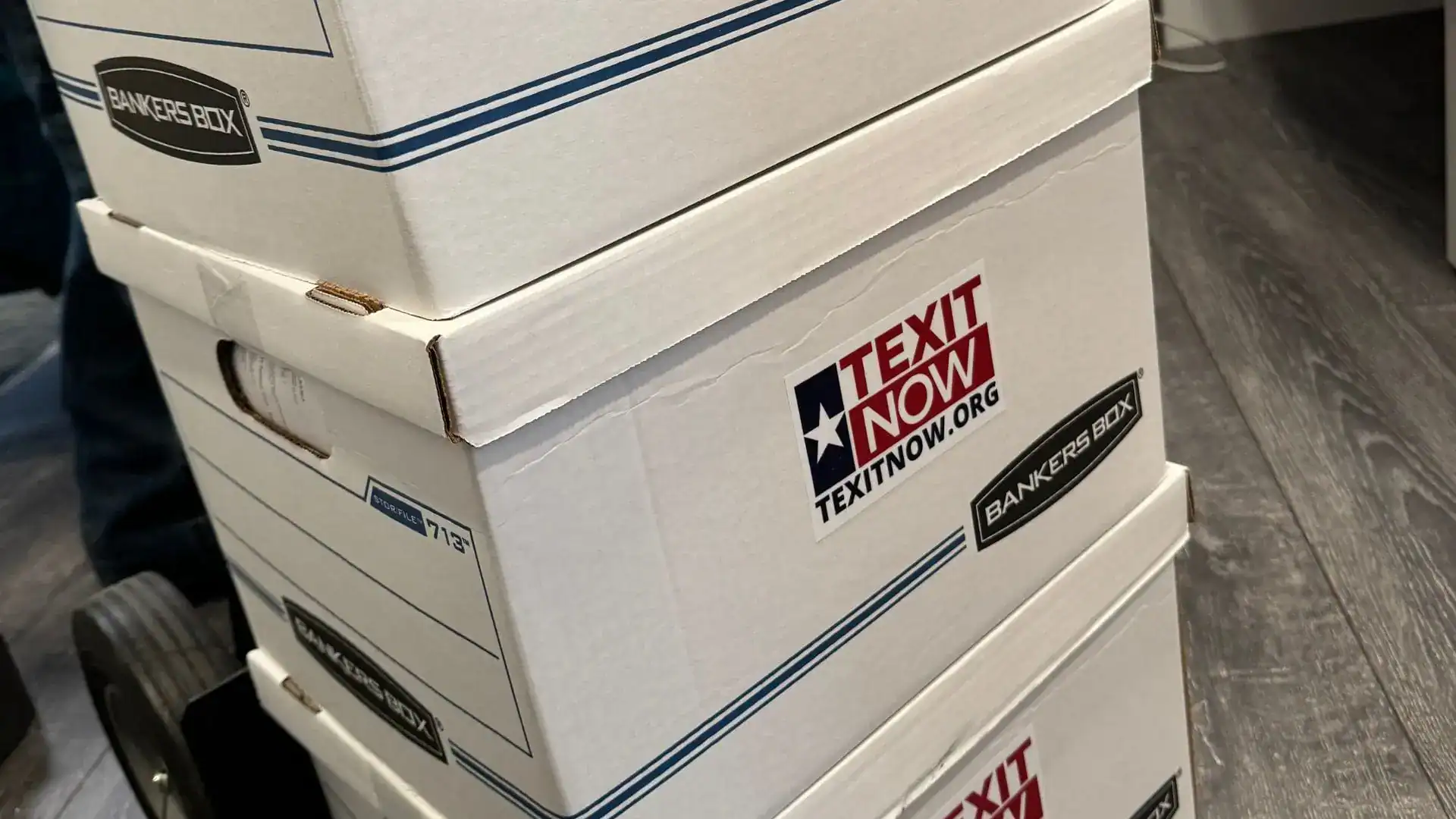 Stacked white file boxes with 'TEXIT Now' stickers on a hand truck.