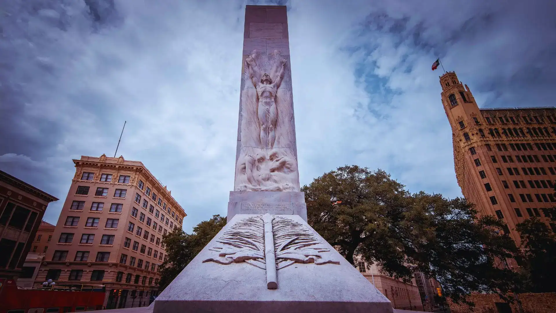 The Alamo Cenotaph in San Antonio seen from below, a carved stone monument against a cloudy sky between downtown buildings.