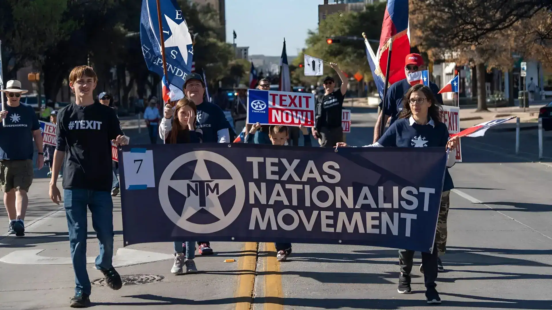 Texas Nationalist Movement supporters march down a street behind a TNM banner, carrying flags and 'TEXIT Now!' signs.