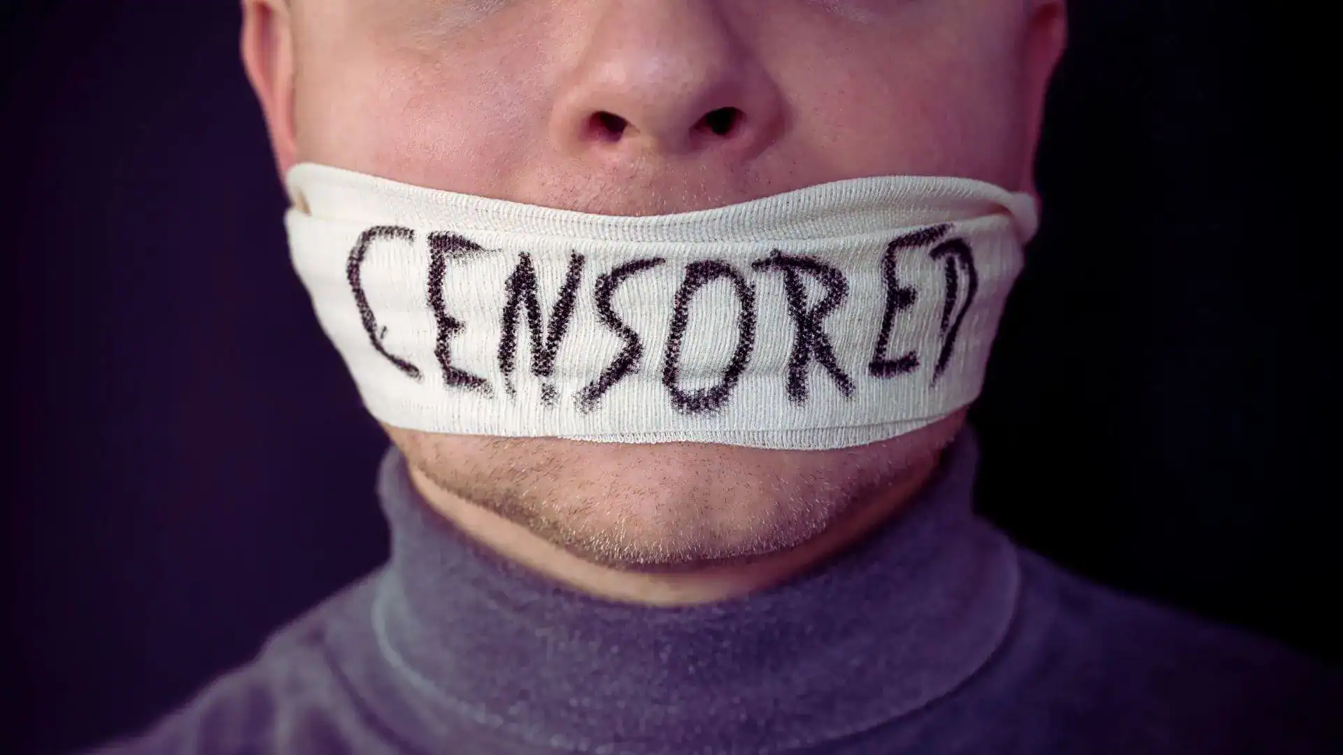 A man with a cloth gag across his mouth marked Censored, wearing a gray turtleneck.