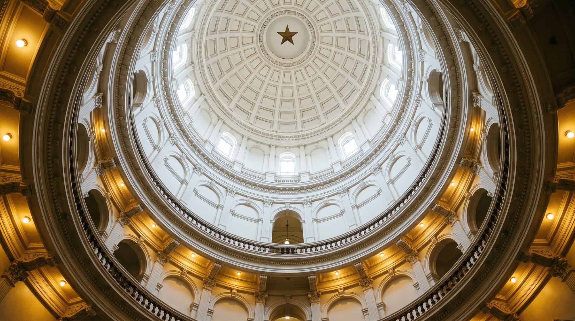 The interior of the Texas State Capitol rotunda, looking up at the ornate white dome with the gold Lone Star at its center, warm architectural light