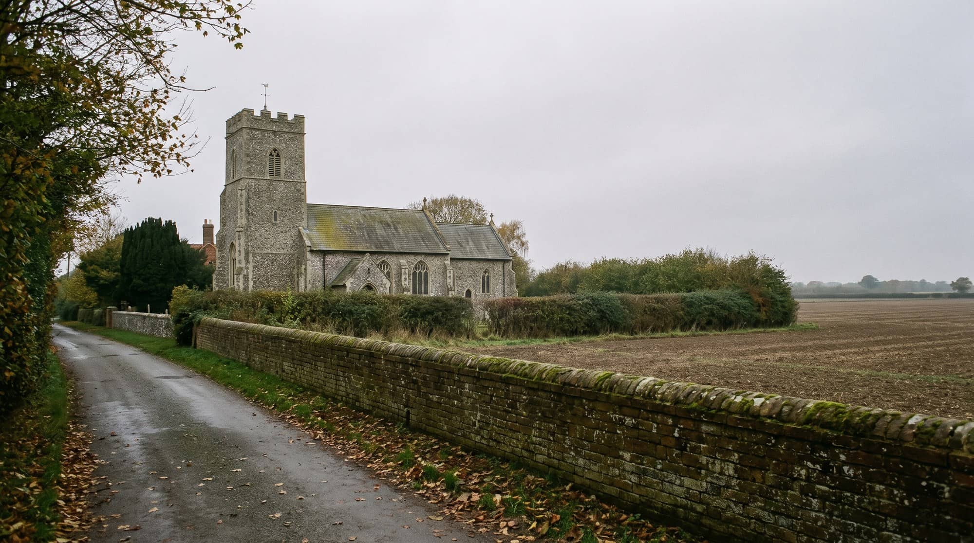 An old flint parish church with a square tower beside a narrow lane and brick wall in rural Suffolk on an overcast autumn day.
