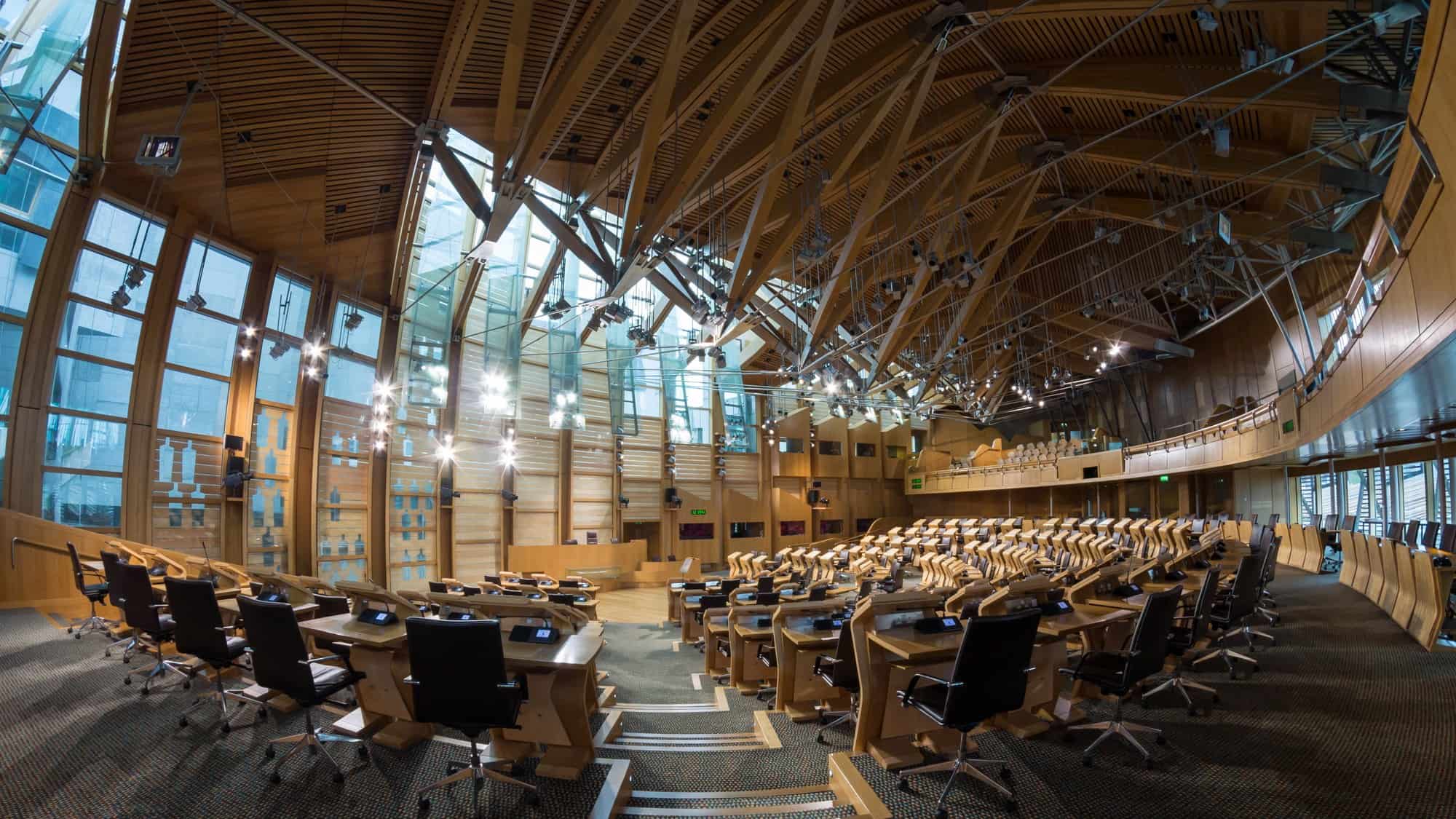 A debating chamber with curved benches and desks, empty, lit from a high glass ceiling.