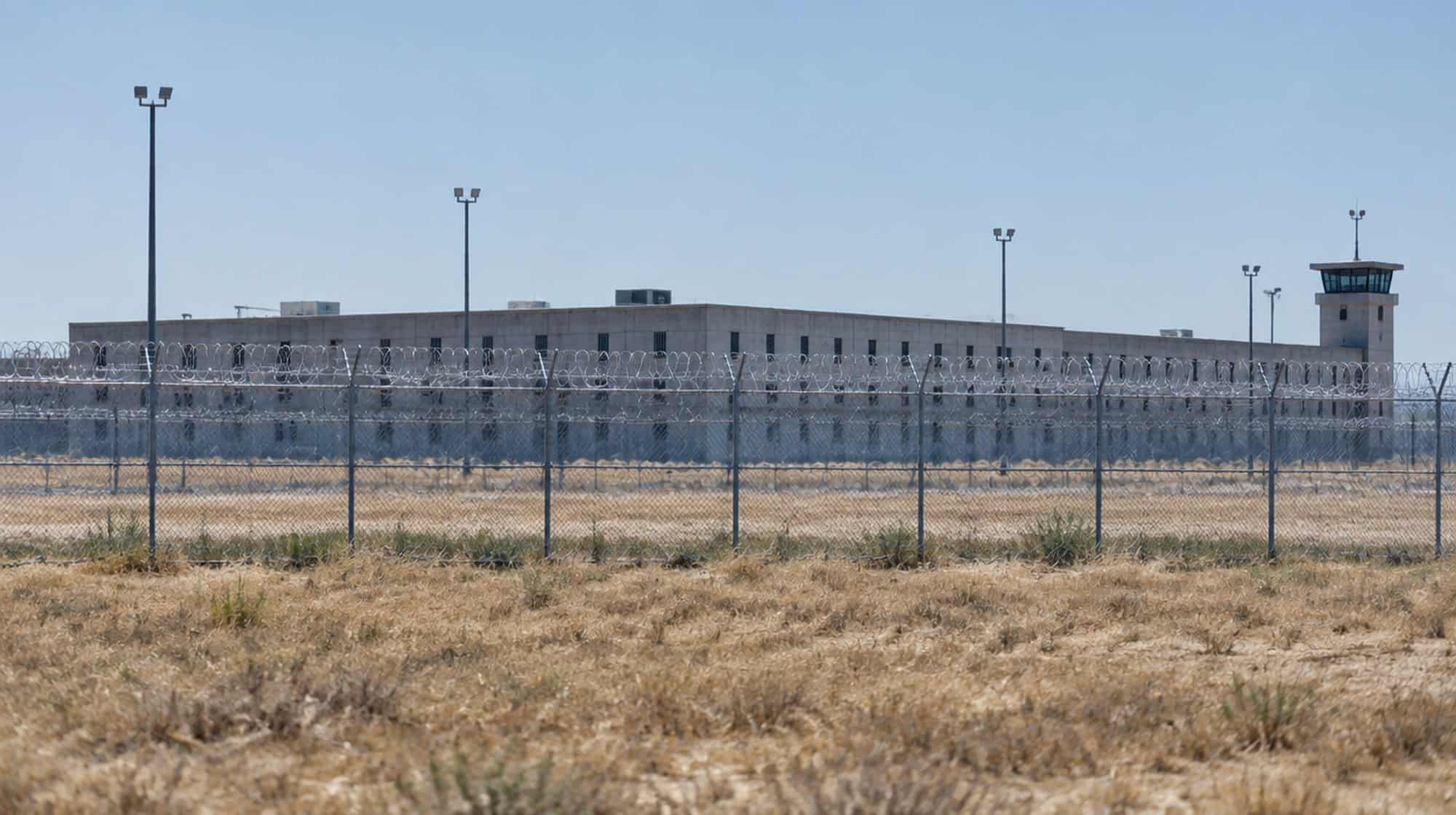 A low concrete prison building behind a chain-link fence topped with razor wire under a hot, hazy sky.