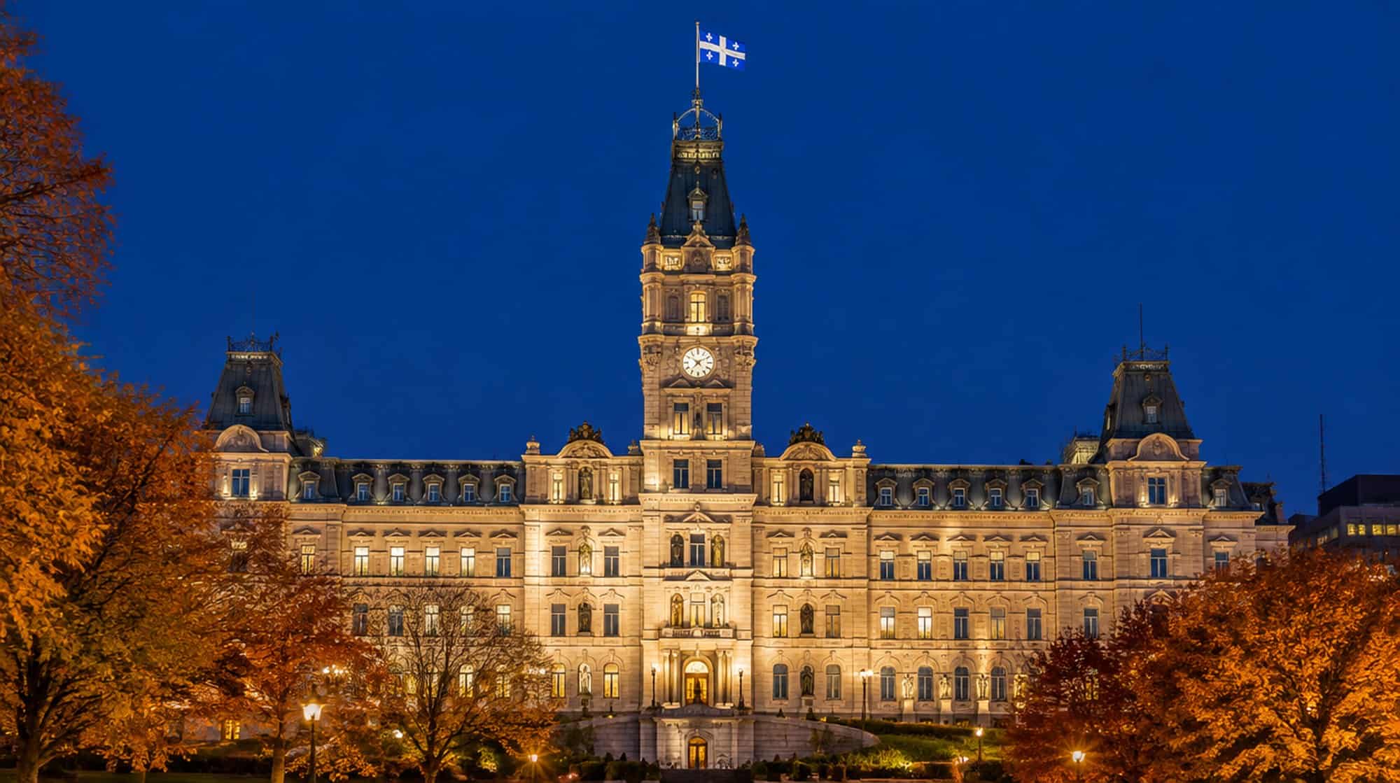 Quebec's Parliament Building in Quebec City lit at night, the Fleurdelisé flying from its clock tower, framed by autumn trees.